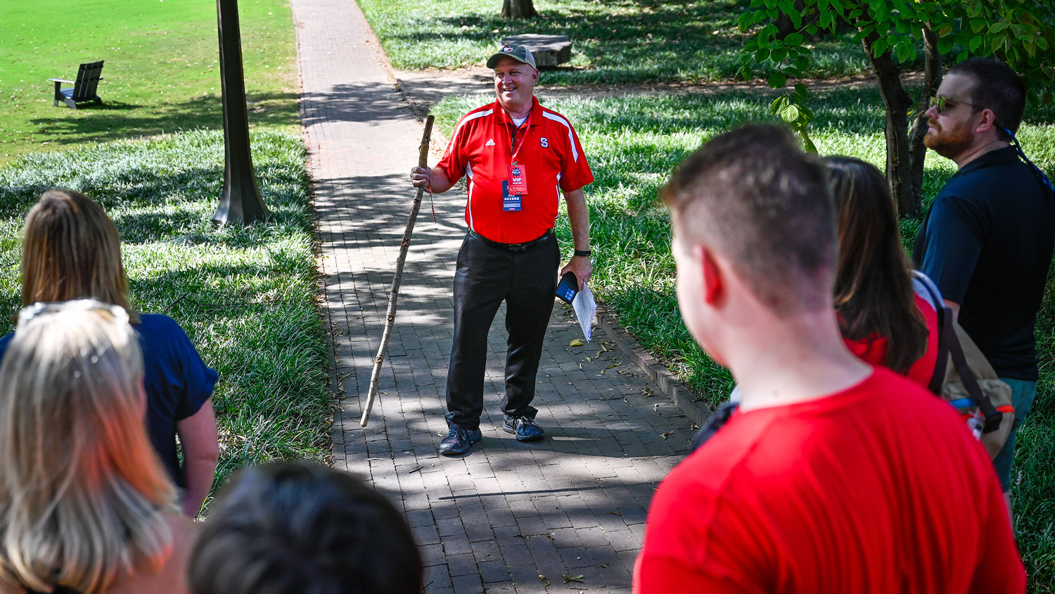 Tim Peeler standing on a brick path and holding a walking stick while speaking to a small crowd of people