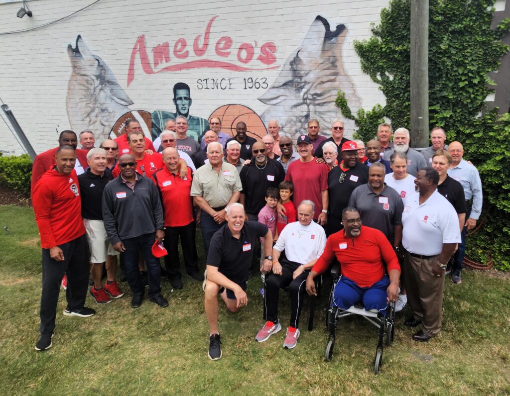 Bill Cowher stands among a large group of former NC State football players, coaches and his other friends from his time at NC State. Behind them is a white wall that reads "Amedeo's since 1963," along with artwork of howling wolf heads and a football and basketball.