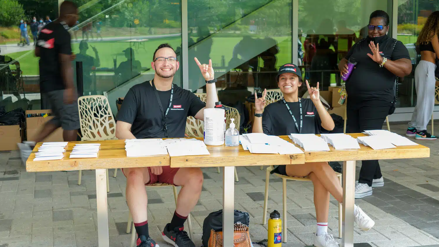 Two students sitting at a registration table make wolfie signs with their hands