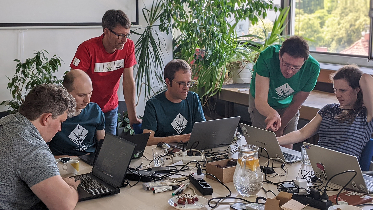 photo shows three people seated at a table working on laptop computers. two additional people are looking over their shoulders. the body language suggests they are working collaboratively on a project.