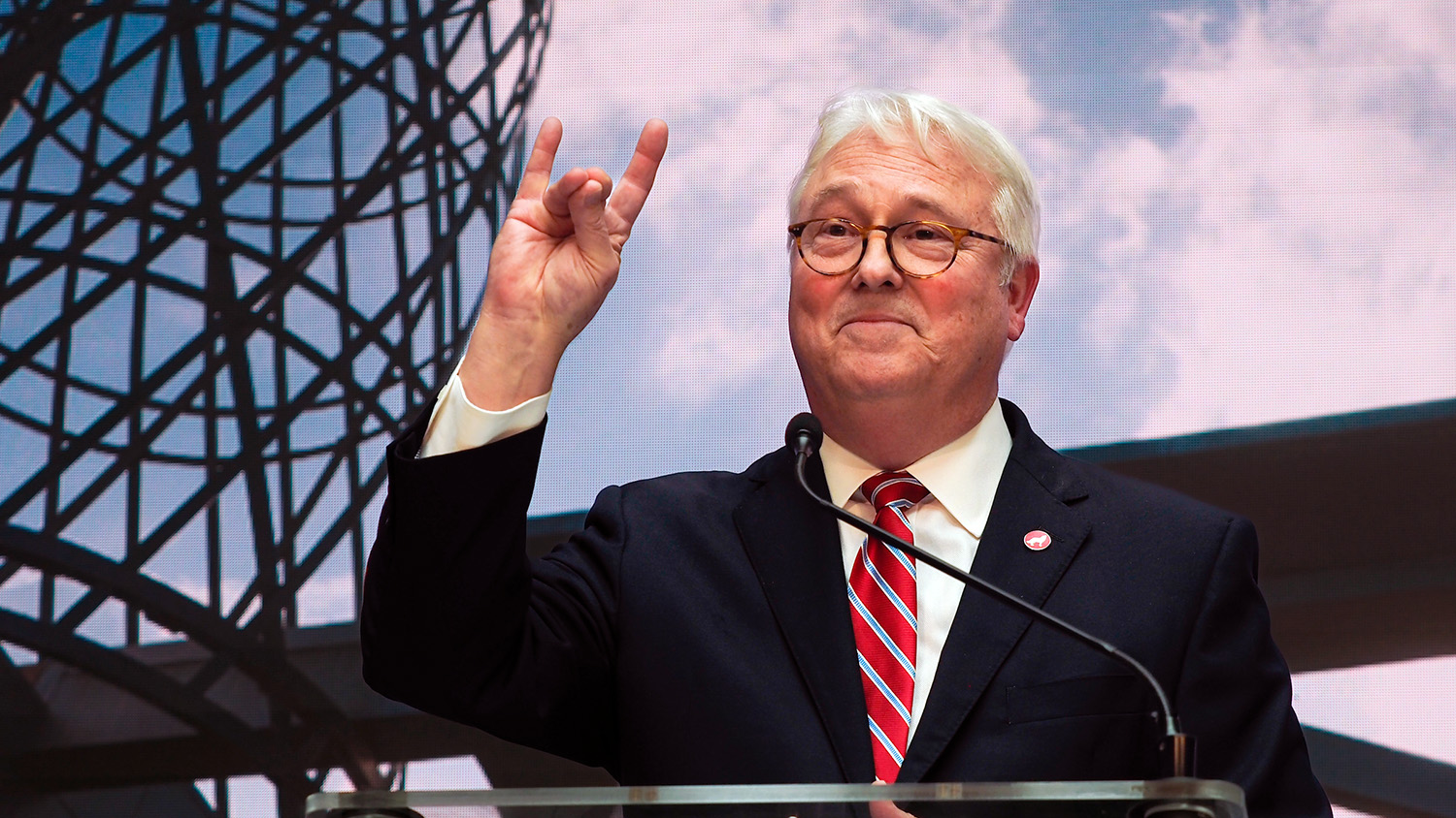 NC State Chancellor Randy Woodson throws up a "wolf" sign as he speaks from a podium.