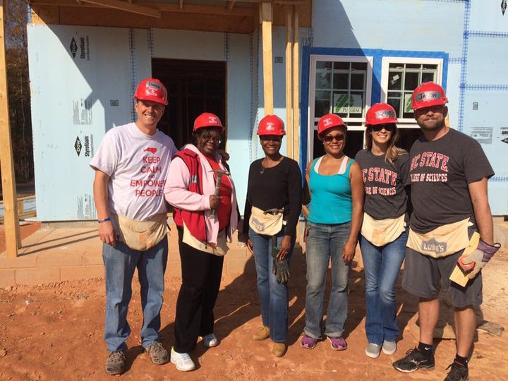 A group of people in red hardhats in front of a half-constructed house for a Build a Block project.