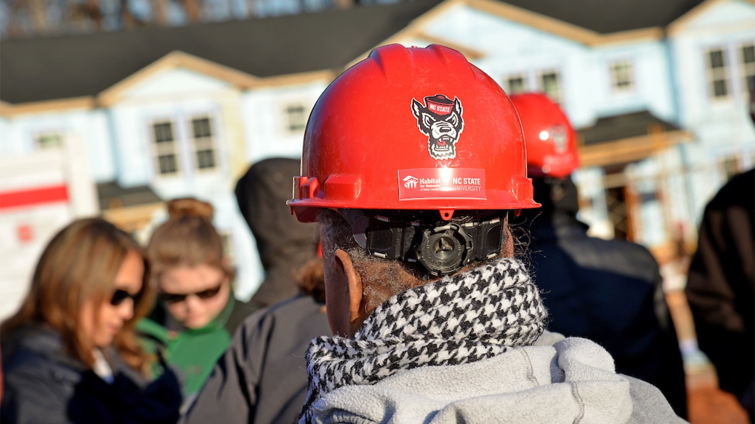 A man wearing a red hardhat with the NC State Wolfpack logo on it, in the middle of a group of volunteers at a Habitat for Humanity build project.