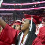 A student takes a selfie in her cap and gown, surrounded by other graduating students, during spring commencement.