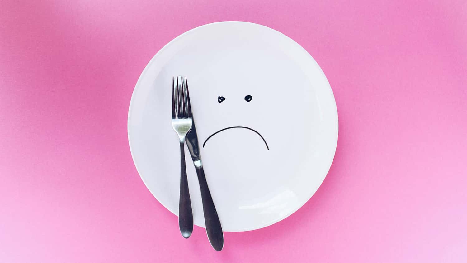 photo shows a plate on a pink background. The plate has a frowny face drawn on it. A knife and fork rest on the plate.