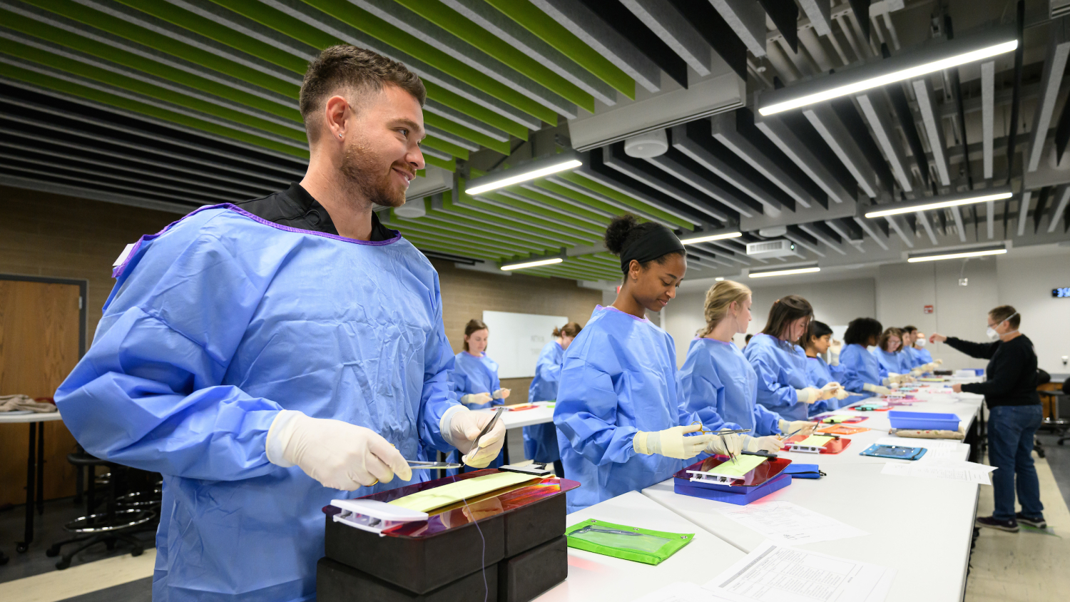 Students in blue surgical gowns and wearing yellow sterile gloves smile while holding suturing needles over fabric models.