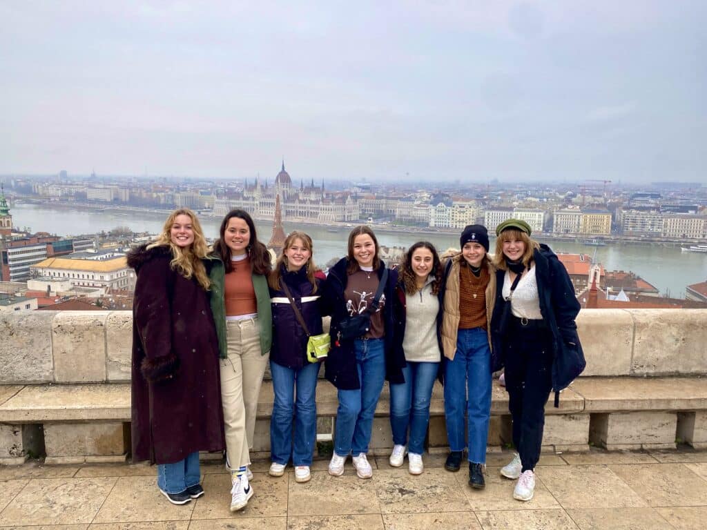 A group of students pose in front of a waterway with a picturesque European skyline in the background.
