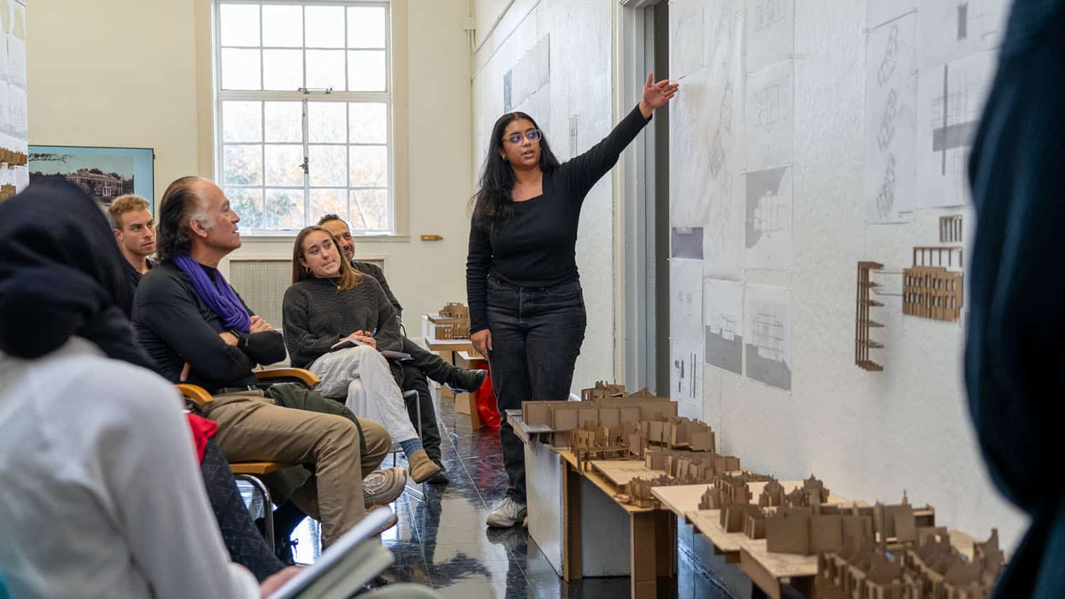A student points to a set of architectural plans on the wall as she presents a project to an audience of peers and faculty.
