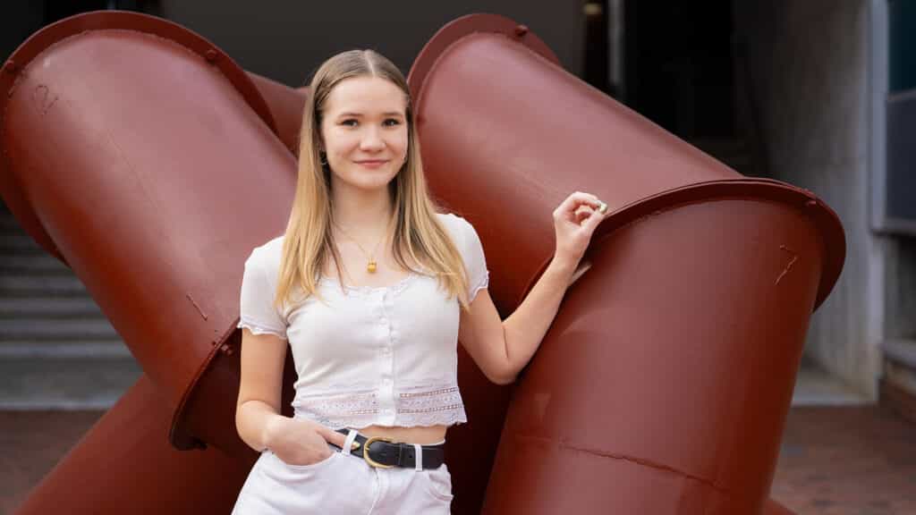 College of Design student Cora Jones poses in front of a large geometric sculpture on campus.