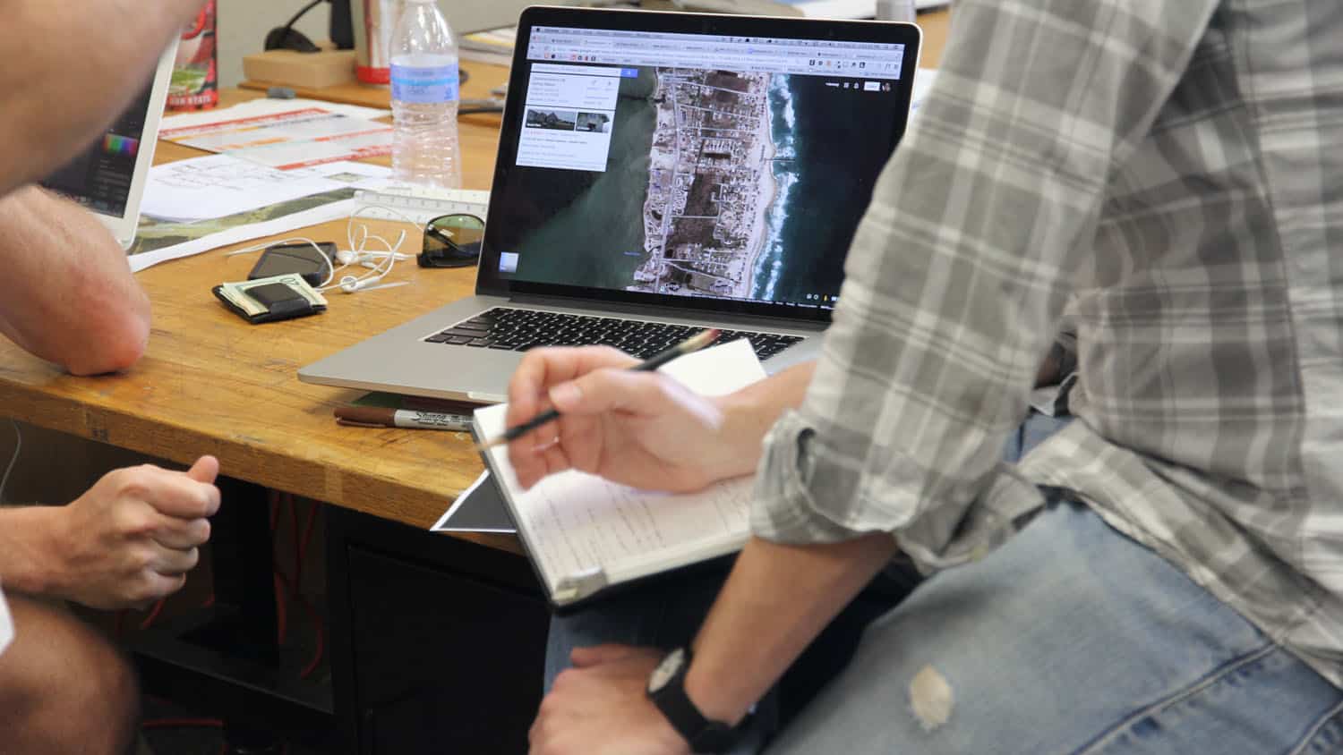 Doctoral design students work over a laptop in a space on campus; the laptop displays an overhead view of a coastal community.