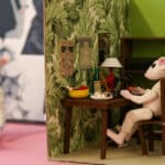 A closeup of a diorama featuring an anthropomorphic mouse seated at a table in a room designed to look like a human house.