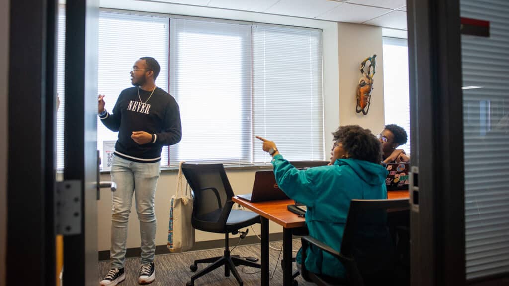A student stands and writes on a whiteboard while two other students sit at a table looking on, inside a conference room in the African American Cultural Center.