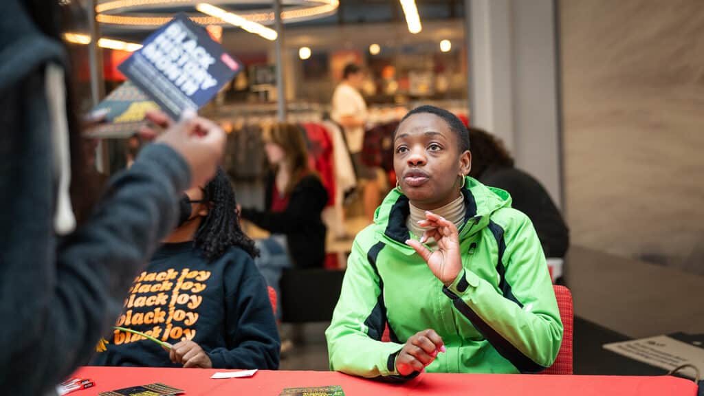A student sits at a table and speaks to another student who is holding a handout that reads "Black History Month" during the kickoff event for Black History Month in the lobby of Talley Student Union.