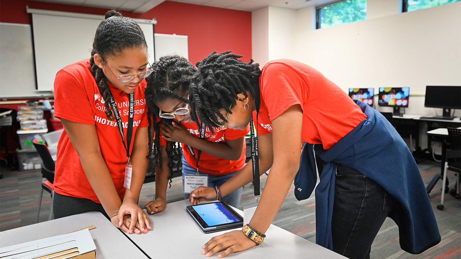Three female students huddle around a tablet on a white table during an IMHOTEP Academy event at The Science House.