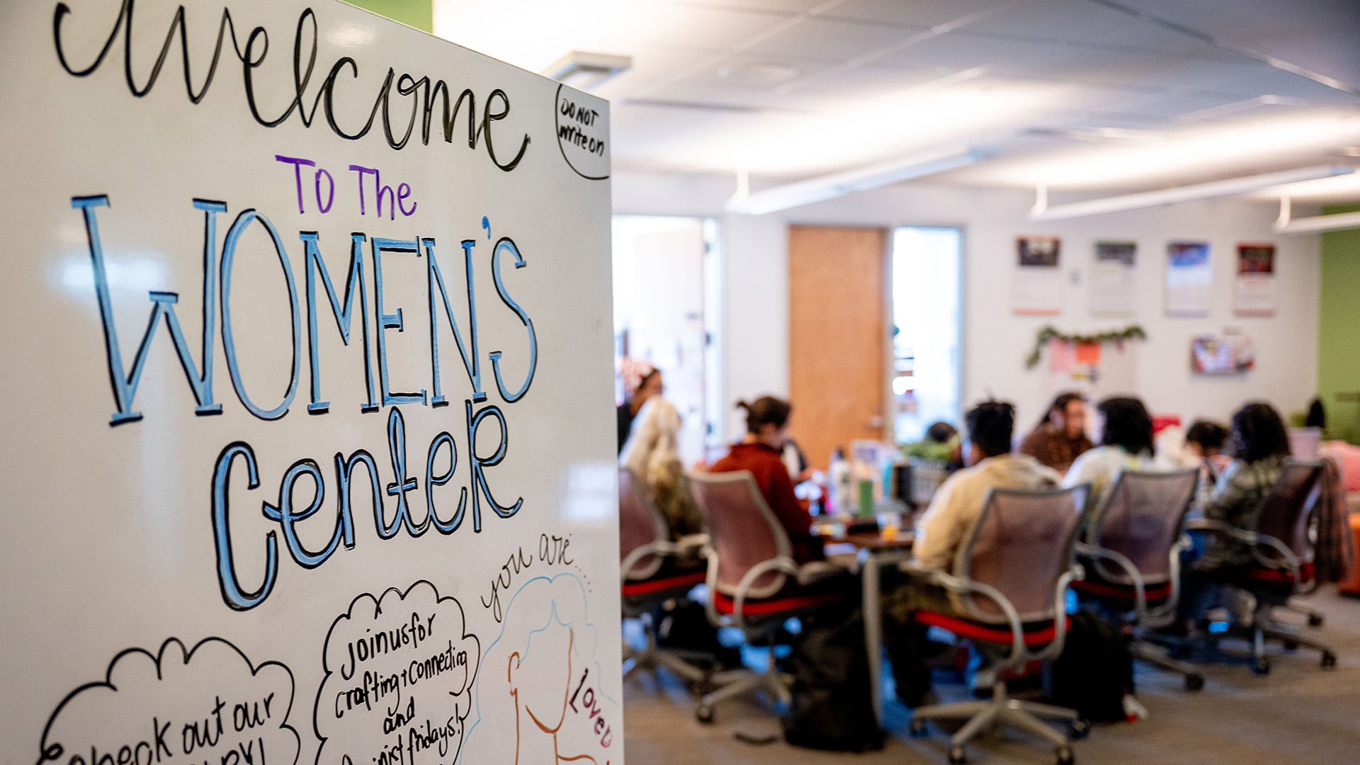 A sign reading "Welcome to the Women's Center", with a group of students sitting around a table in the background.