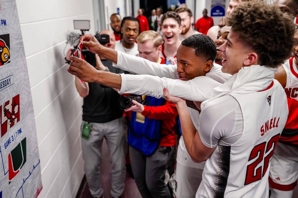 NC State men's basketball players hold up KJ Keatts as he places the NC State logo on a bracket board showing the team in the Sweet 16. 
