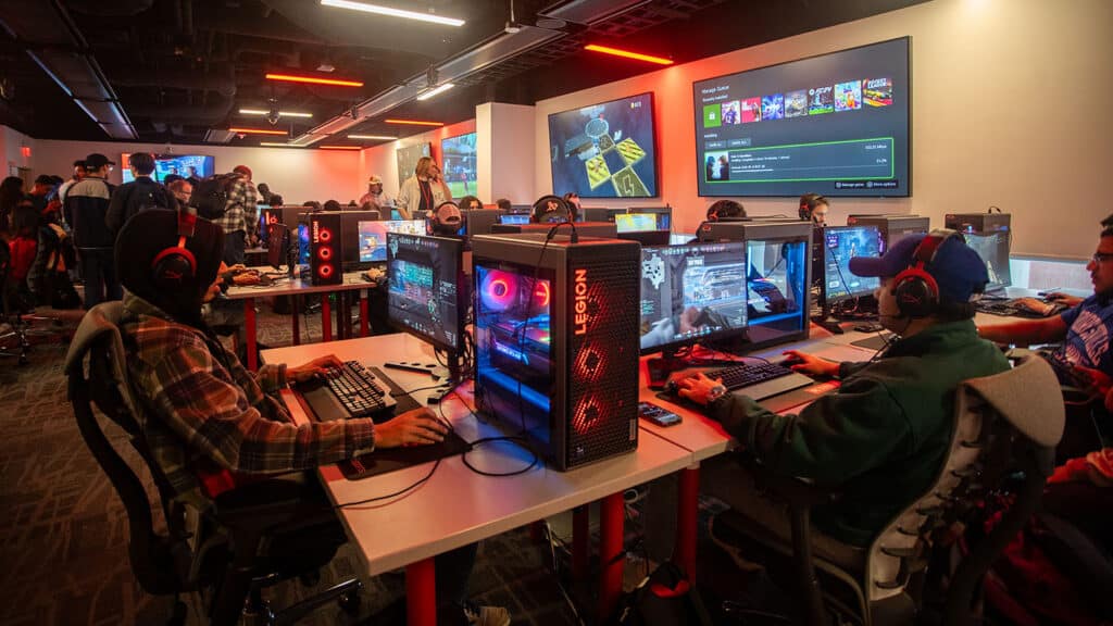 A wide-angle view of students playing video games at computer stations in the NC State Gaming and Esports Lab, with orange lights illuminating the room.