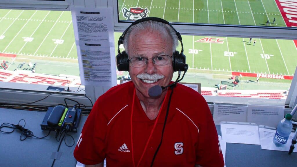 Gary Hahn wearing a headset in the broadcast booth, with the football field of Carter-Finley Stadium behind him.