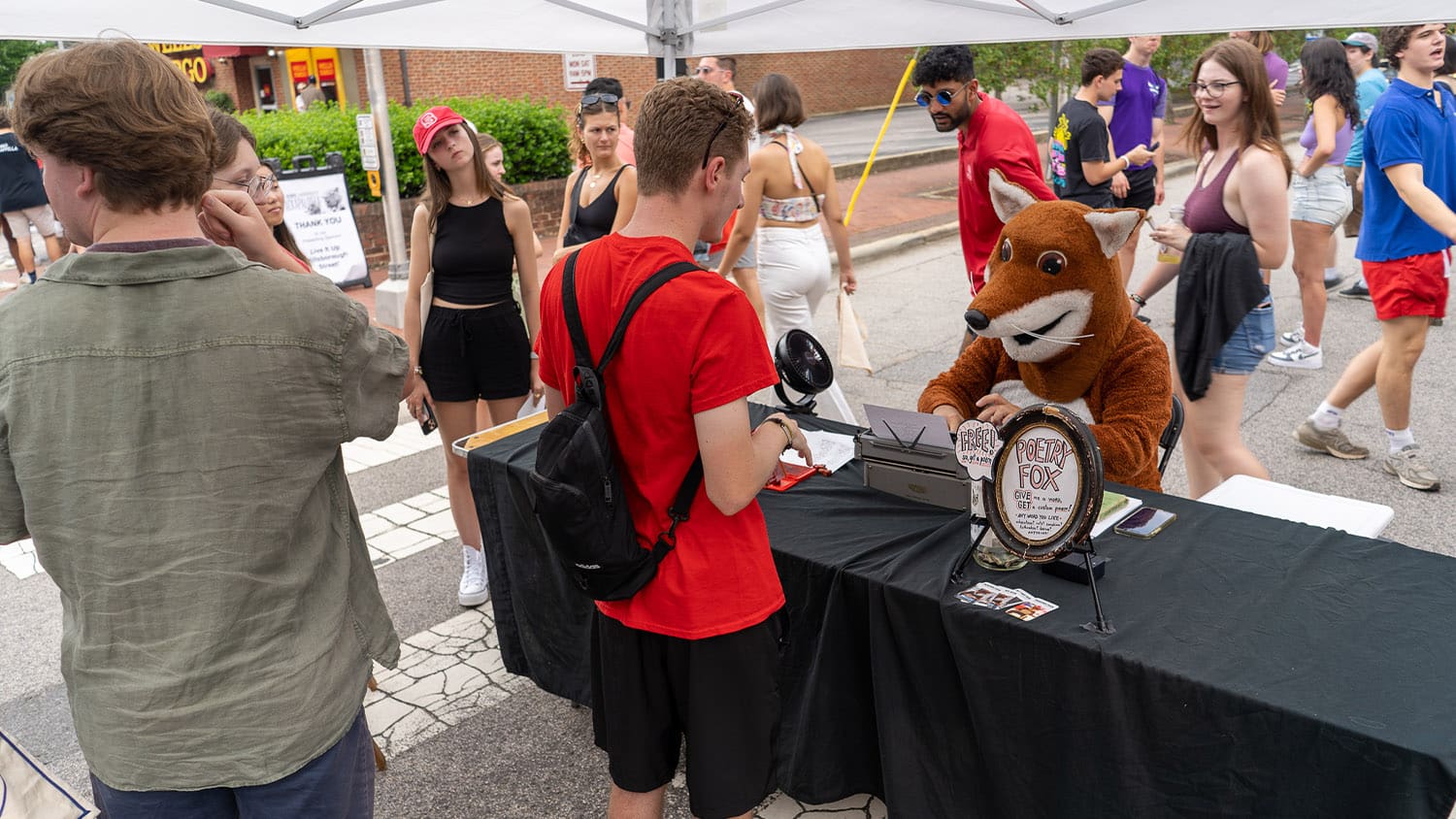 Chris Vitiello (the Poetry Fox) sits at a table under a tent and types poems for students during Packapalooza.