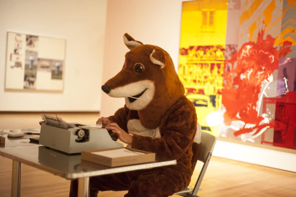 Vitiello in the Poetry Fox costume, sitting at a table and typing on his typewriter in the middle of a museum gallery