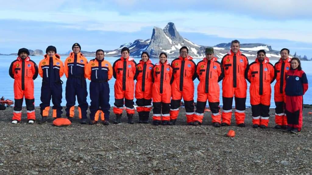 A team of researchers pose in Antarctica with water behind them and rocks rising in the distance.