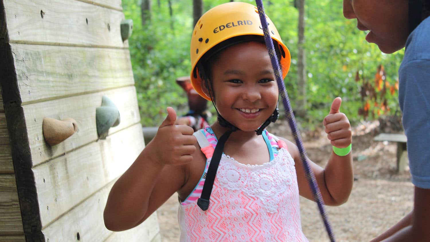 A young camper gives two thumbs up as she prepares to climb a climbing wall.