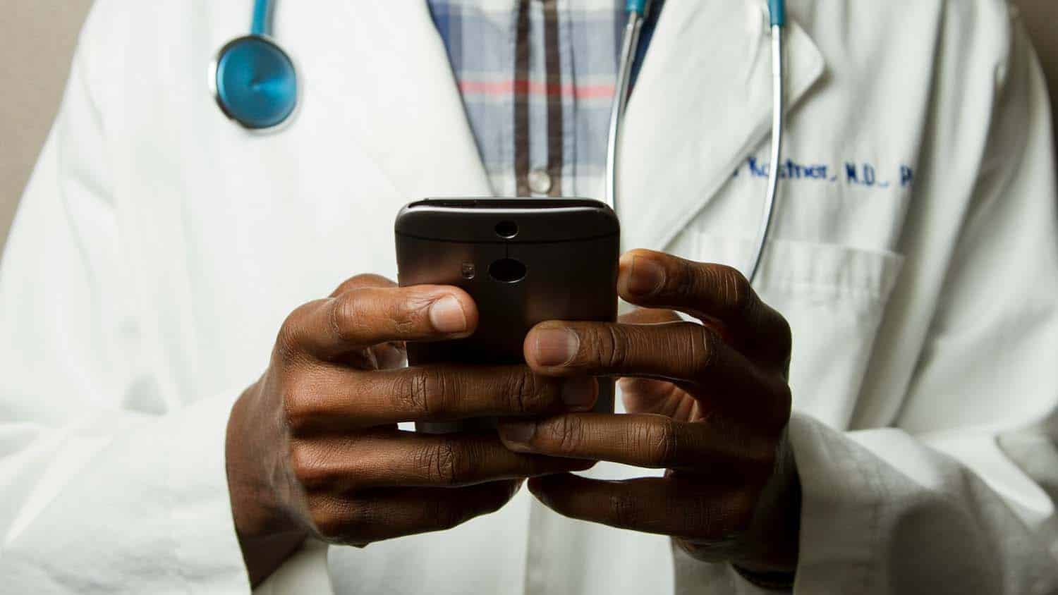 photo shows a black person in white labcoat marked with the letters MD, PhD. The person has a stethoscope around their neck and is holding a smartphone.