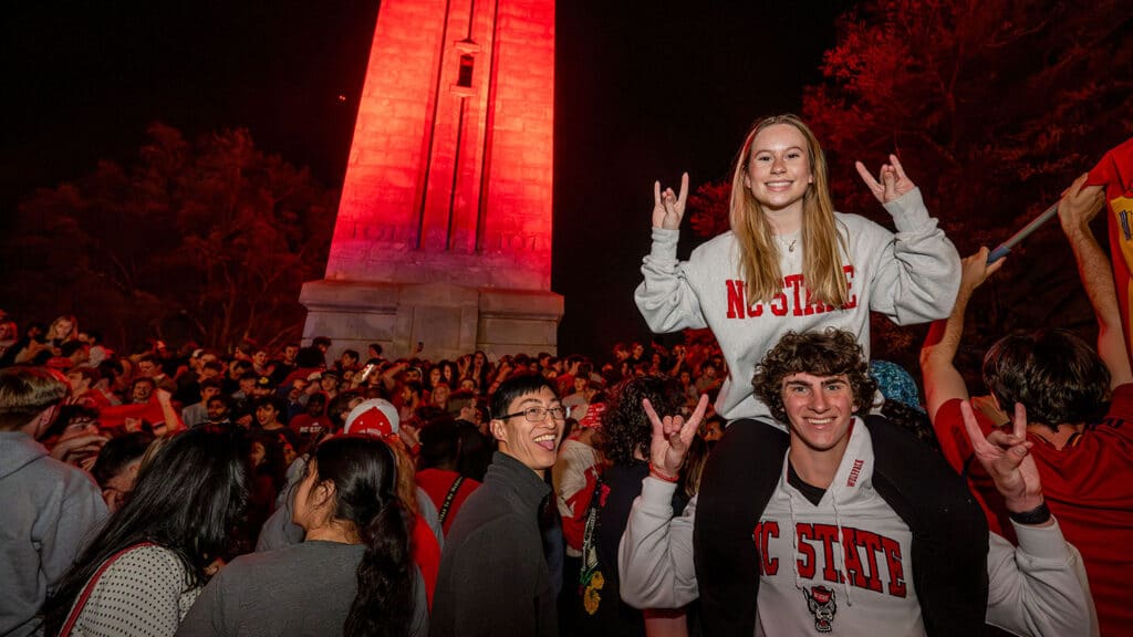 A large crowd of students and fans at the NC State Belltower at night. The tower is illuminated in red.