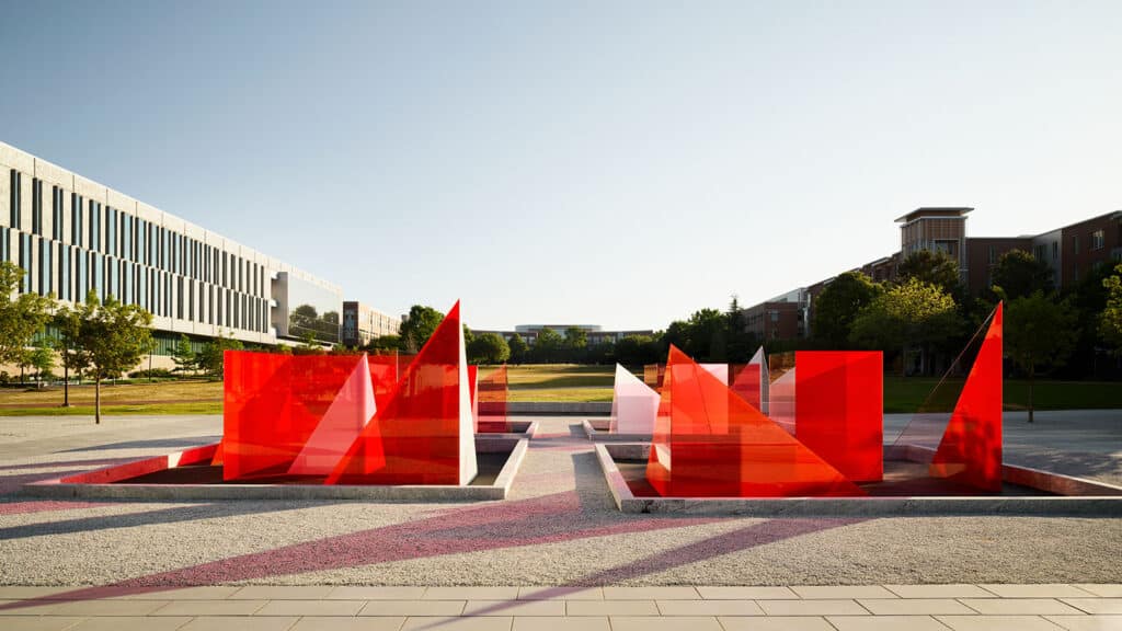 An ground-level view of the Reds and Whites art installation by artist Larry Bell, showing glass red and white glass pieces clustered in a central plaza between stands of trees outside the James B. Hunt Jr. Library on Centennial Campus.