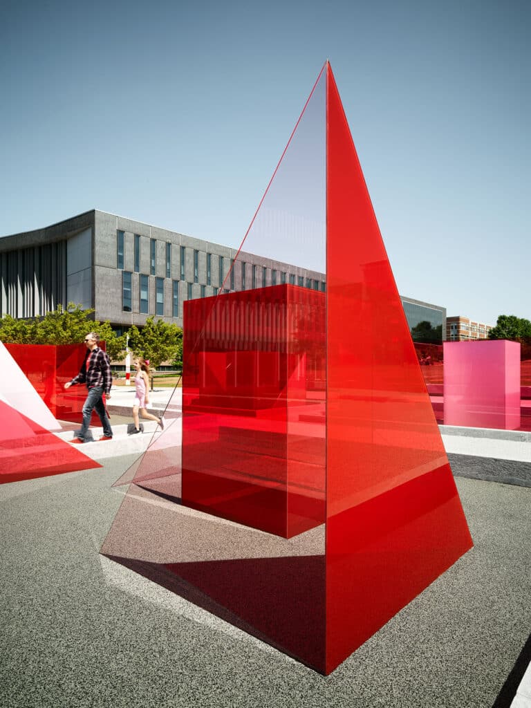 A view of one of the component pieces of the Reds and Whites art installation by artist Larry Bell, showing a pyramidal glass red and white glass piece clustered in a central plaza between stands of trees outside the James B. Hunt Jr. Library on Centennial Campus.