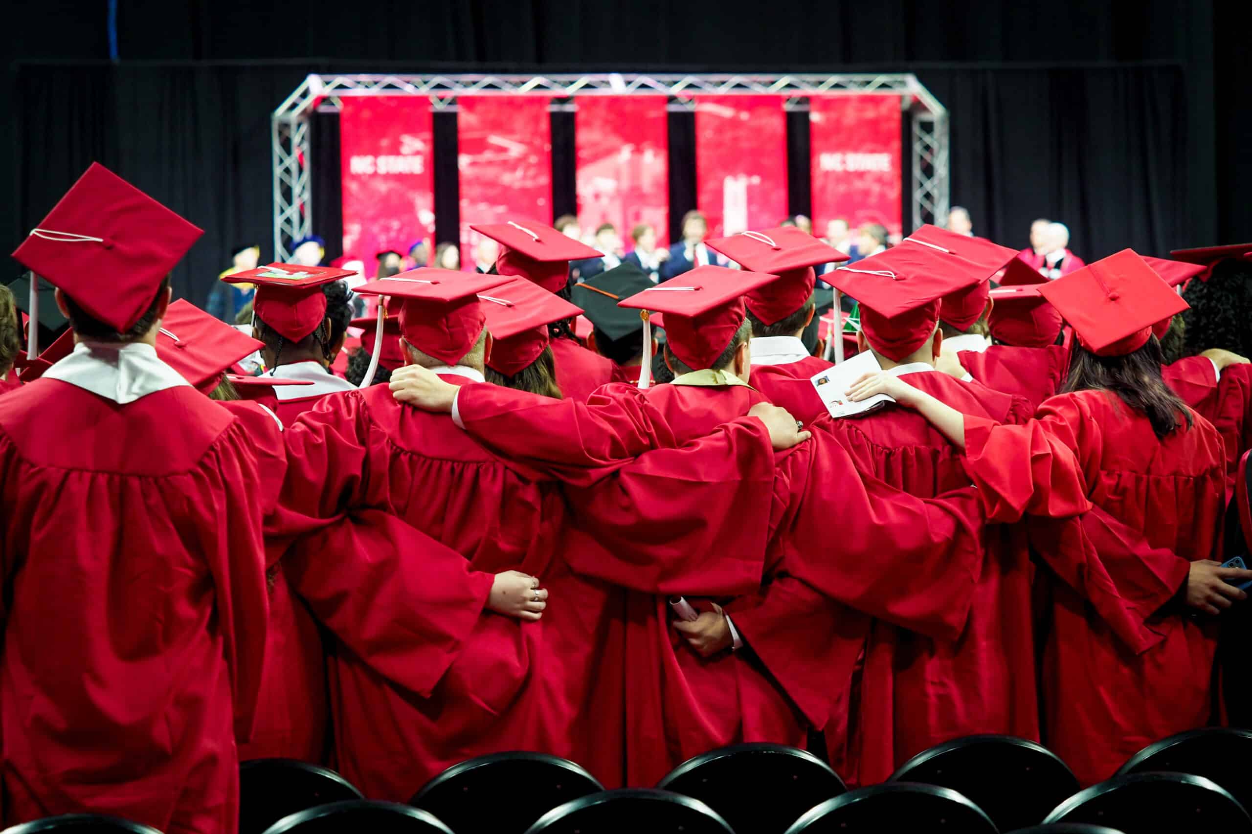 Students in their red graduation robes embrace and face the stage in PNC Arena