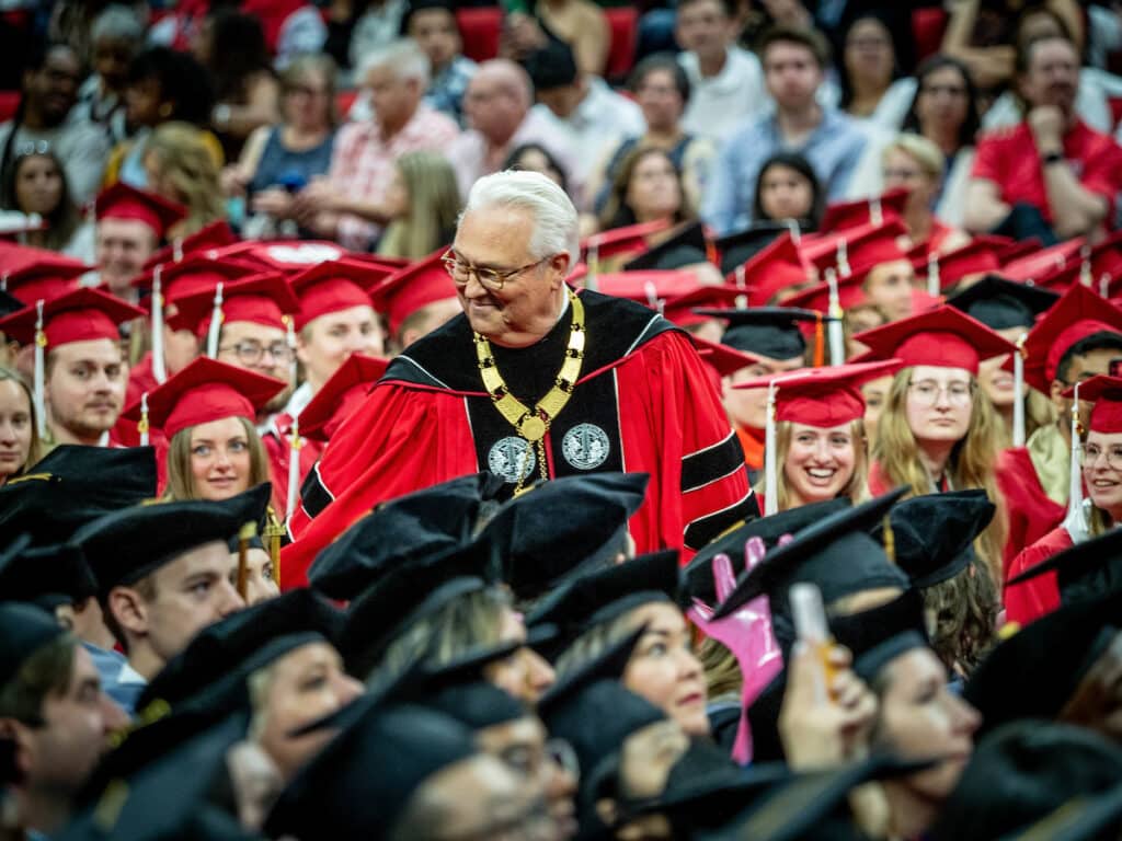 Chancellor Woodson standing among a large group of seated NC State students in red and and black graduation hats and robes. 