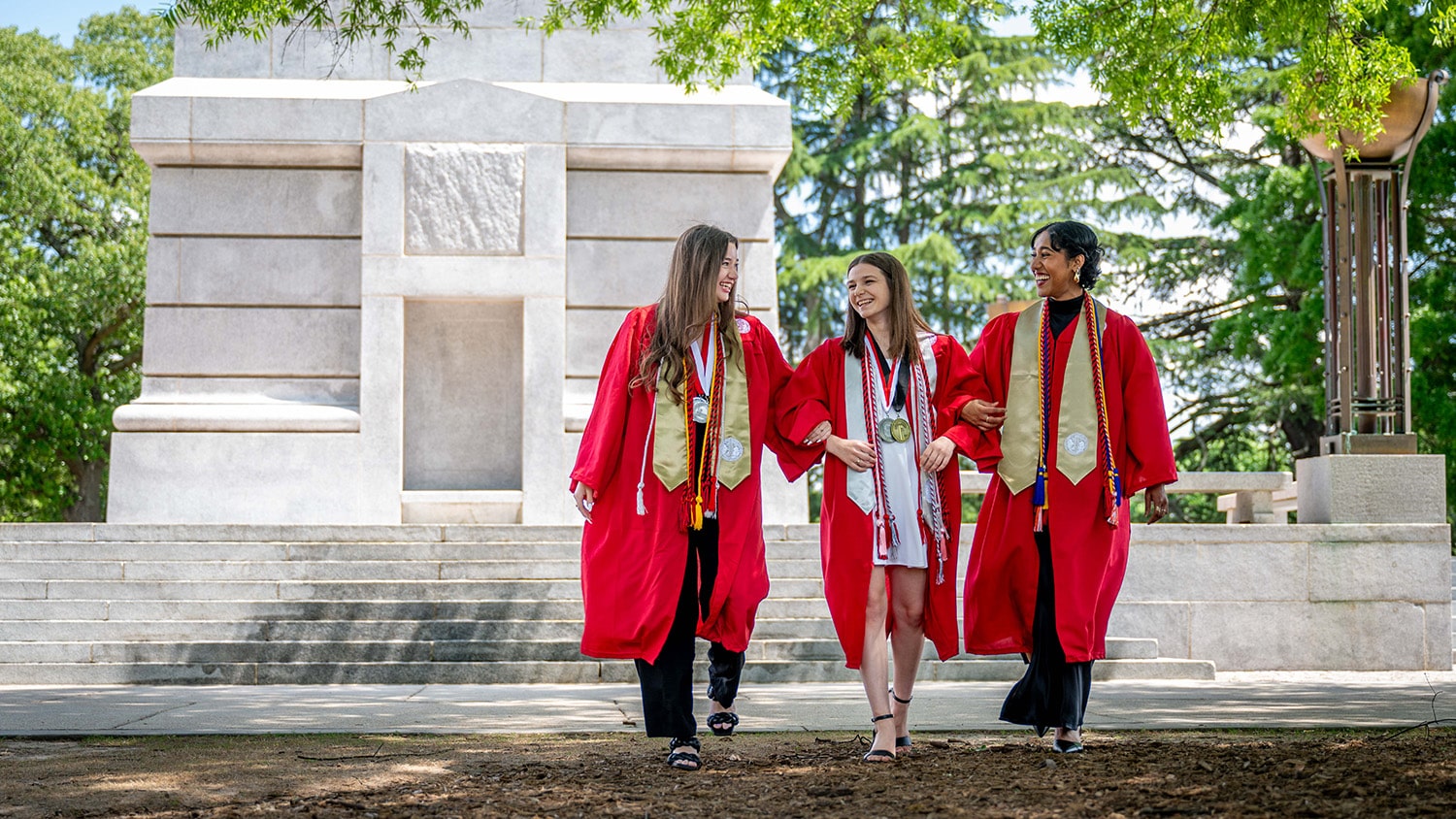 Three chancellor's aides walk arm-in-arm away from the Memorial Belltower wearing their graduation gowns.