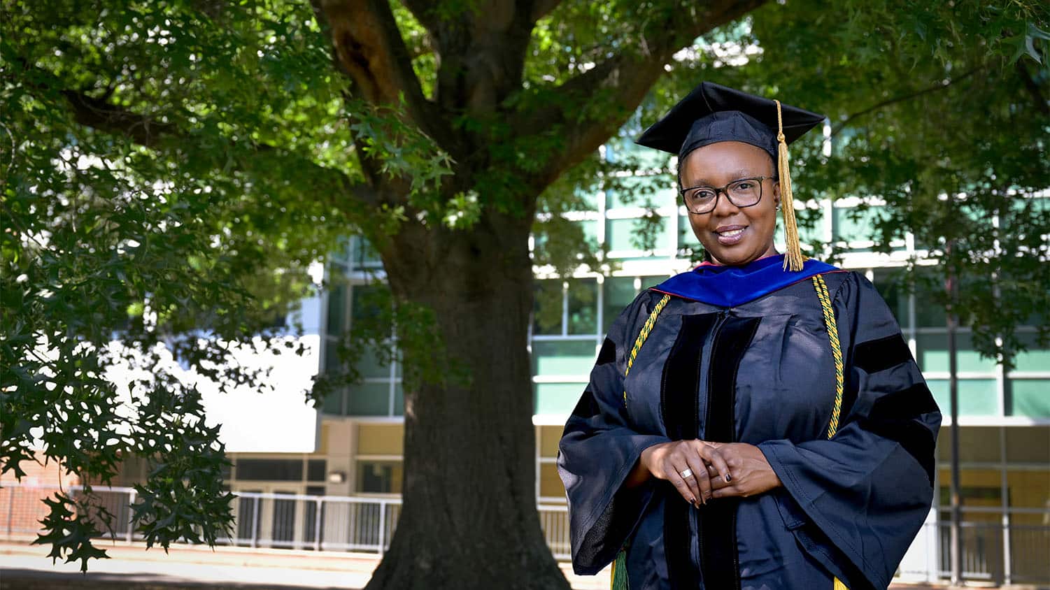 A doctoral graduate student in her cap and gown stands before an large oak tree.