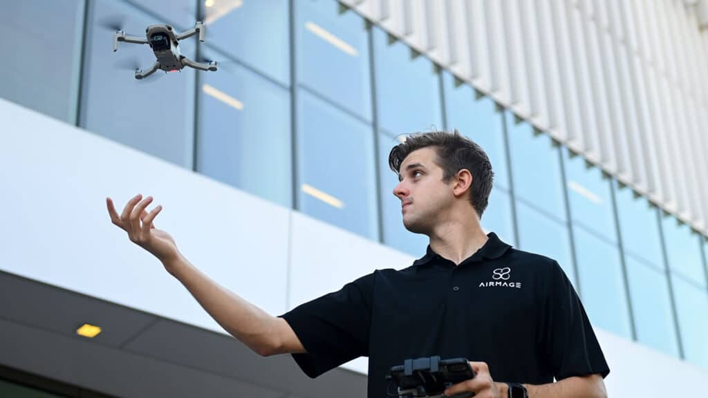 A student stands in front of a building with a remote in his hand releasing a drone into the air.