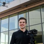 A student poses with a camera while a drone hovers over his shoulder.