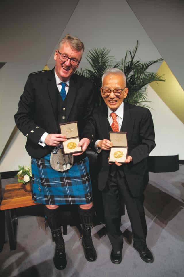 David MacMillan, wearing a kilt, holds his Nobel Prize medal next to a colleague