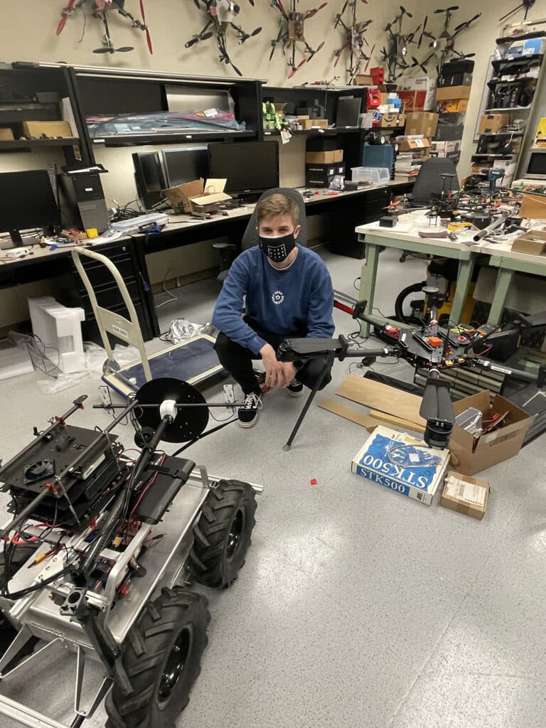 A student, wearing a mask, poses in a workshop surrounded by remote-controlled vehicles.