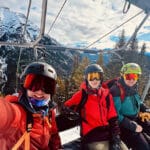 A selfie-style photo shows three males in ski outfits smiling while sitting in a ski lift.