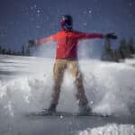 A photo of a snowboarder in orange jacket posing in motion, arms outstretched, as his snowboard throws a shower of snow around him.