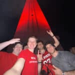 A group of students hold the wolf-ear pose while standing in front of NC State's Memorial Belltower, which is lighted red for a special occasion.