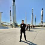 A student in suit and sunglasses poses, arms crossed, in front of an exhibit of various space rockets.