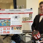 A student smiles while standing in front of a poster showing a research proposal for an AI drone.