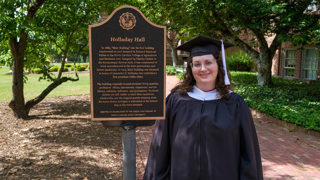 Thompson in her black master's graduation robes, standing next to a sign for Holladay Hall