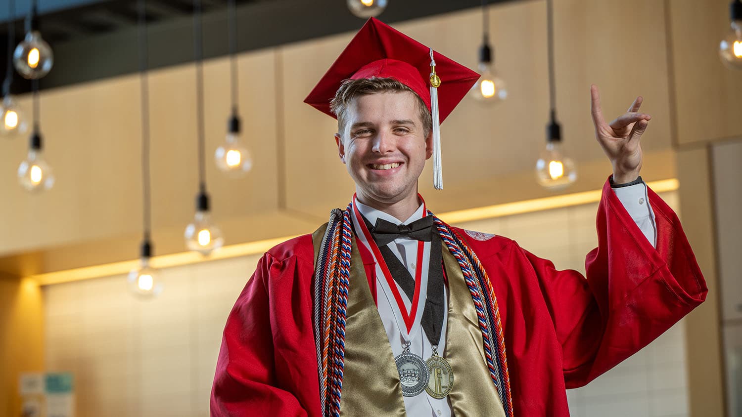 2024 NC State graduate Shaun Deardorff holds the wolf-ear pose in the Raleigh Founded location on Centennial Campus, with lightbulbs shining in the background.