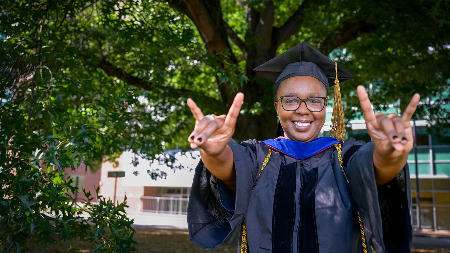 Portrait of Wizaso Munthali wearing her cap and gown and holding up two wolfie hand gestures, standing beneath a large oak tree.