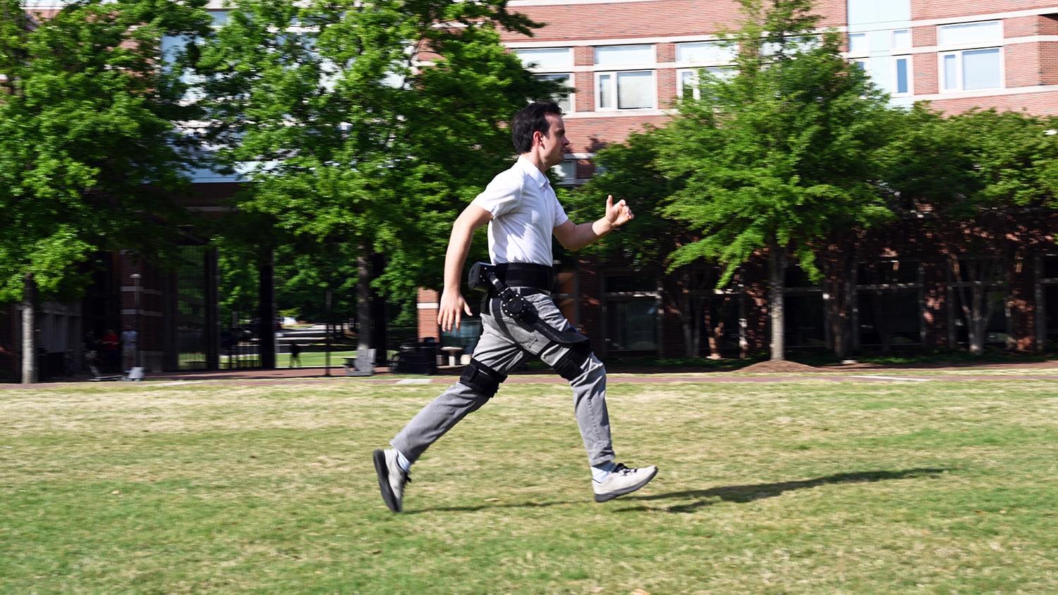 a man runs across a green lawn wearing a robotic exoskeleton that attaches to his waist, hips and knees