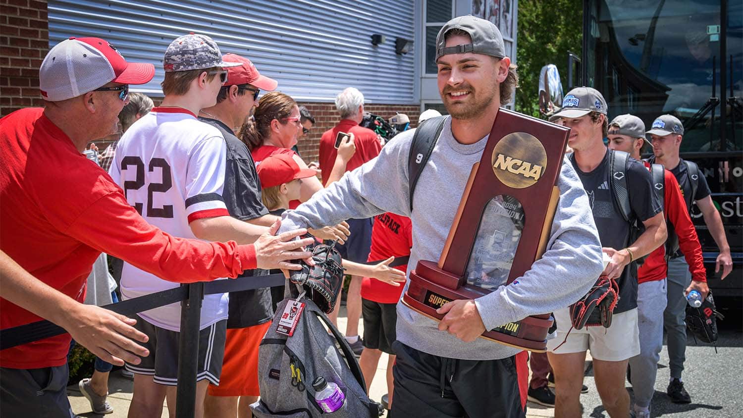 A baseball player in street clothes greets a long line of fans while holding an NCAA trophy
