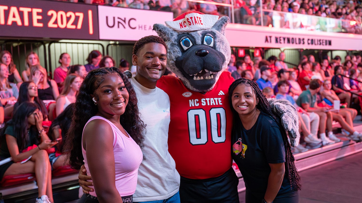 Three students pose for a photo with Mr. Wuf in front of a crowd of other students on bleachers during New Student Convocation.