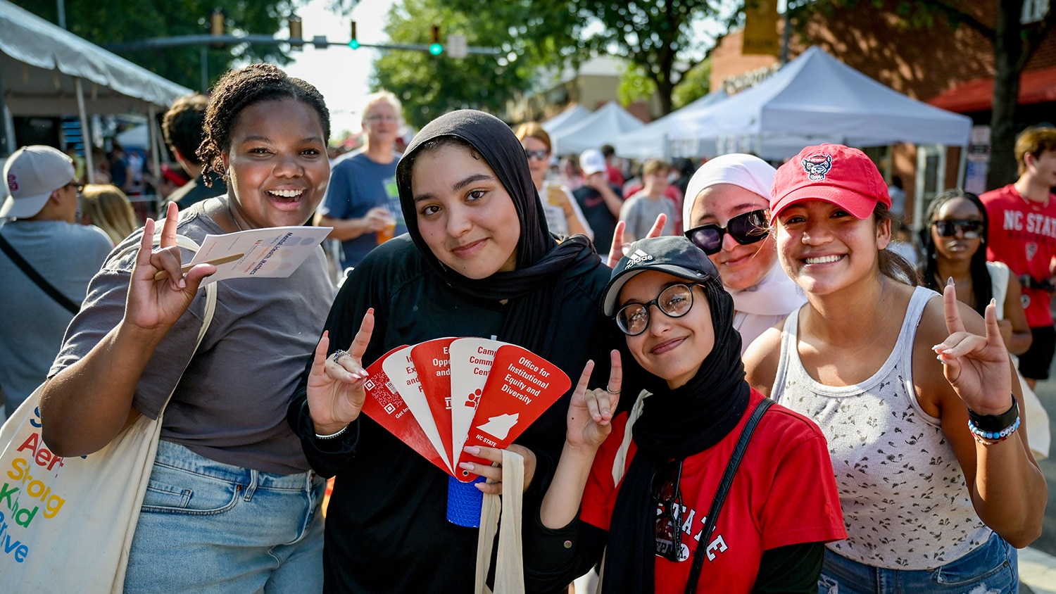 Four students smile and hold up the wolfie hand gesture with a large crowd and tents behind them during Packapalooza.